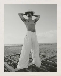 Shirtless man wearing wide-leg white trousers and a wide-brimmed hat standing on a wooden frame structure near the coast.