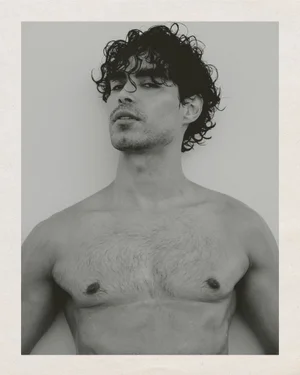 Black and white portrait of a shirtless man with curly dark hair looking upward against a plain background