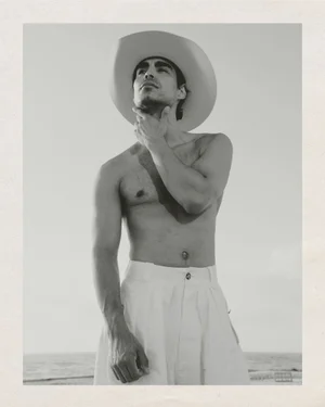 Shirtless man wearing a cowboy hat and light trousers posing outdoors near the ocean in a black and white portrait