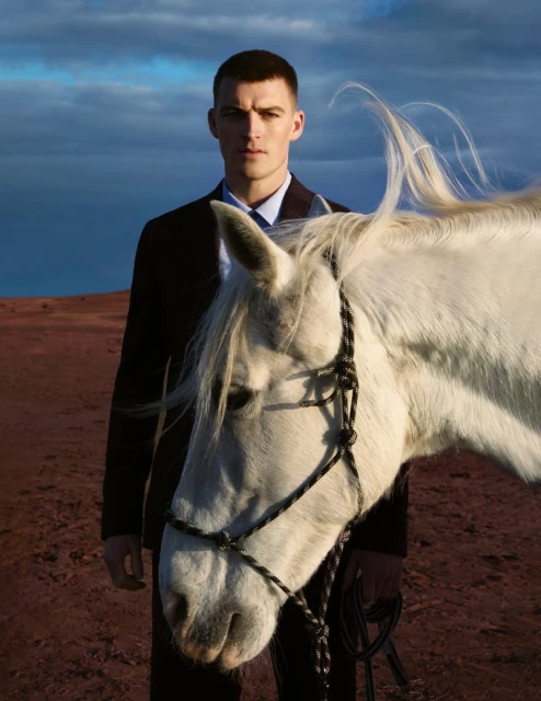 A white horse stands next to a man in a suit outdoors against a partly cloudy sky, creating a striking and unique visual contrast.