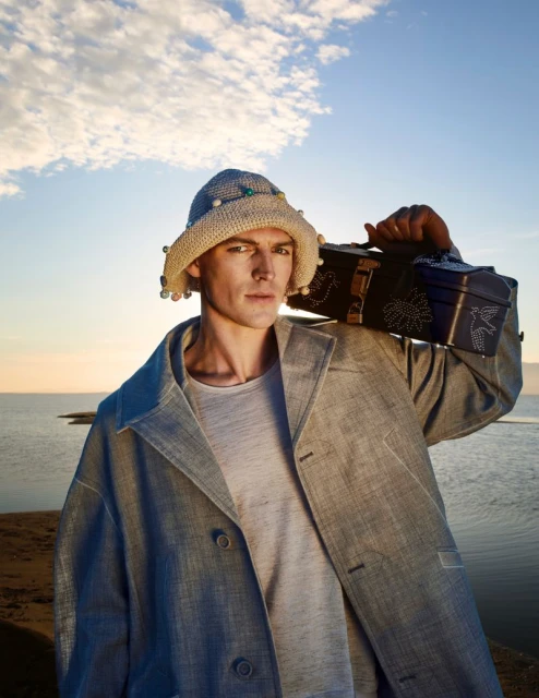 A stylish adult male poses outdoors wearing a beige bucket hat, grey t-shirt, and a jacket, carrying a decorated box on his shoulder.