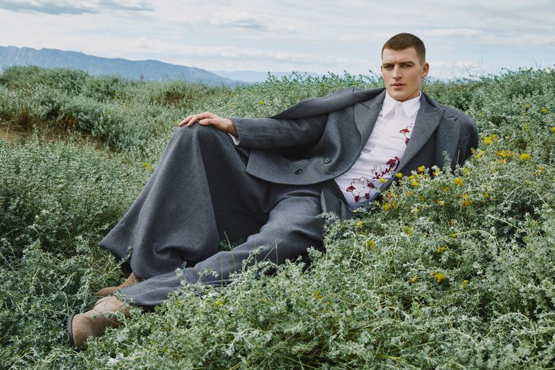Male model in a gray suit and floral shirt lies in a field of green plants with mountains visible in the background under a cloudy sky.