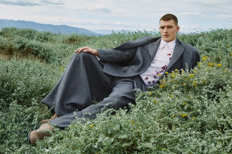 Male model in a gray suit and floral shirt lies in a field of green plants with mountains visible in the background under a cloudy sky.