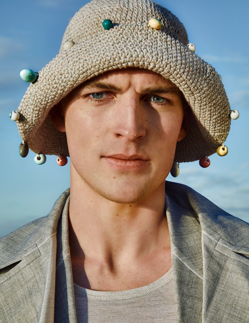 Close-up of adult male wearing a bucket hat decorated with beads, tan jacket, against a bright blue sky, outdoor fashion shot.