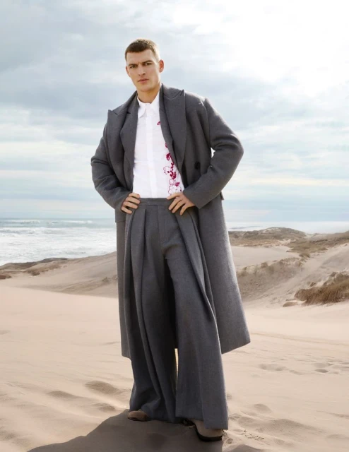 Man poses in oversized grey coat and pleated pants on sand dunes near the ocean under a cloudy sky, fashion editorial photography.