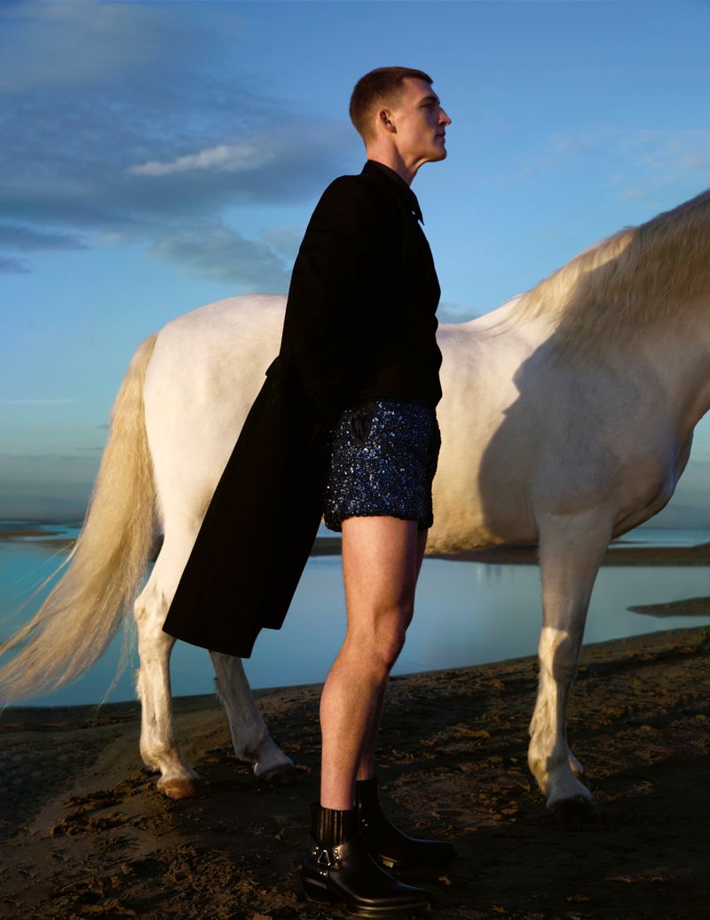 Adult male posing with a white horse on a beach with black coat and blue sequined shorts. Clear blue sky is in the background.
