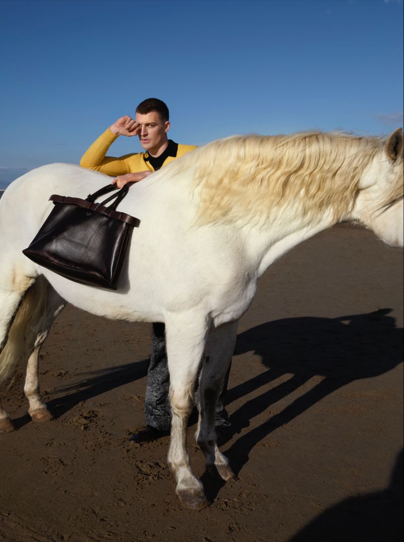 Man in yellow top on a beach with white horse and black bag, posing in the sun with bright blue sky in the background.