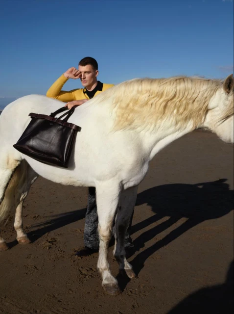 Man in yellow top on a beach with white horse and black bag, posing in the sun with bright blue sky in the background.