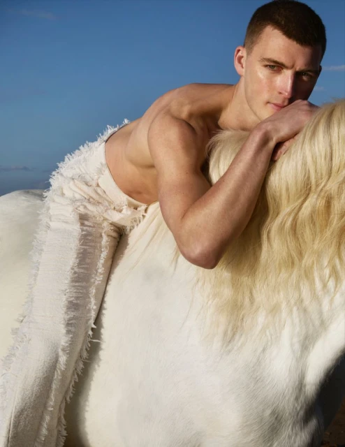 Nude male model poses atop a white horse under a clear blue sky in a fashion editorial. He wears fringed white trousers.