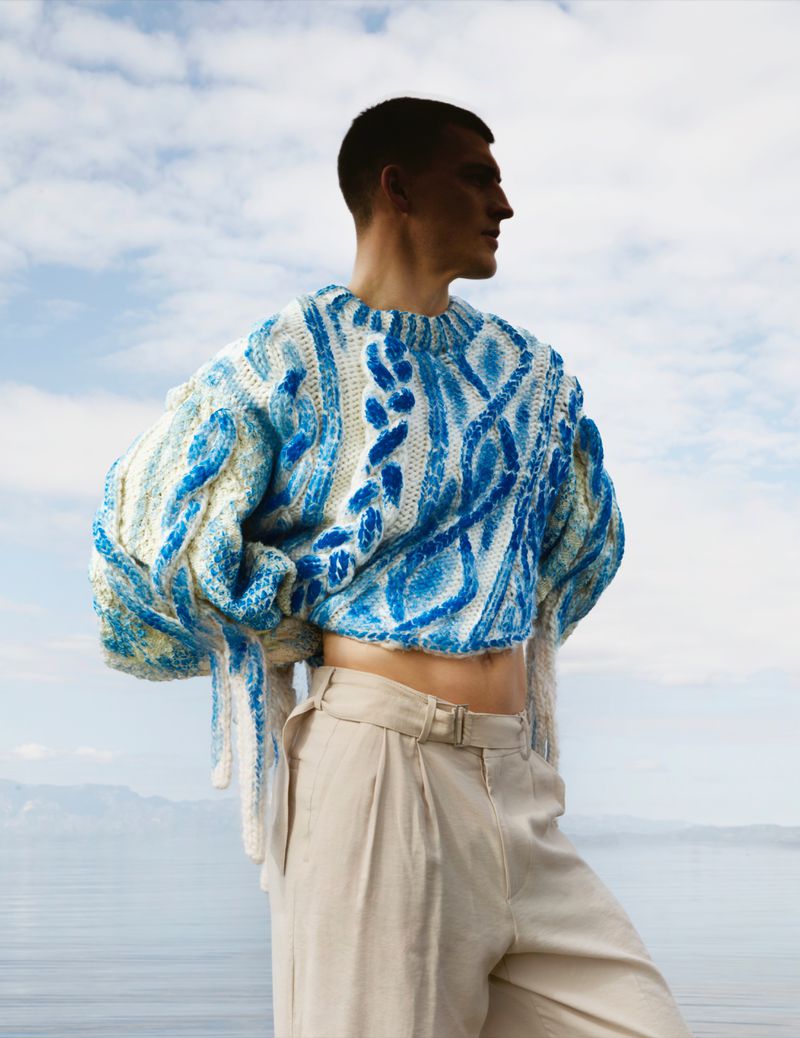 Model wearing a blue and white patterned cable-knit sweater and beige trousers against a cloudy sky and open water background.