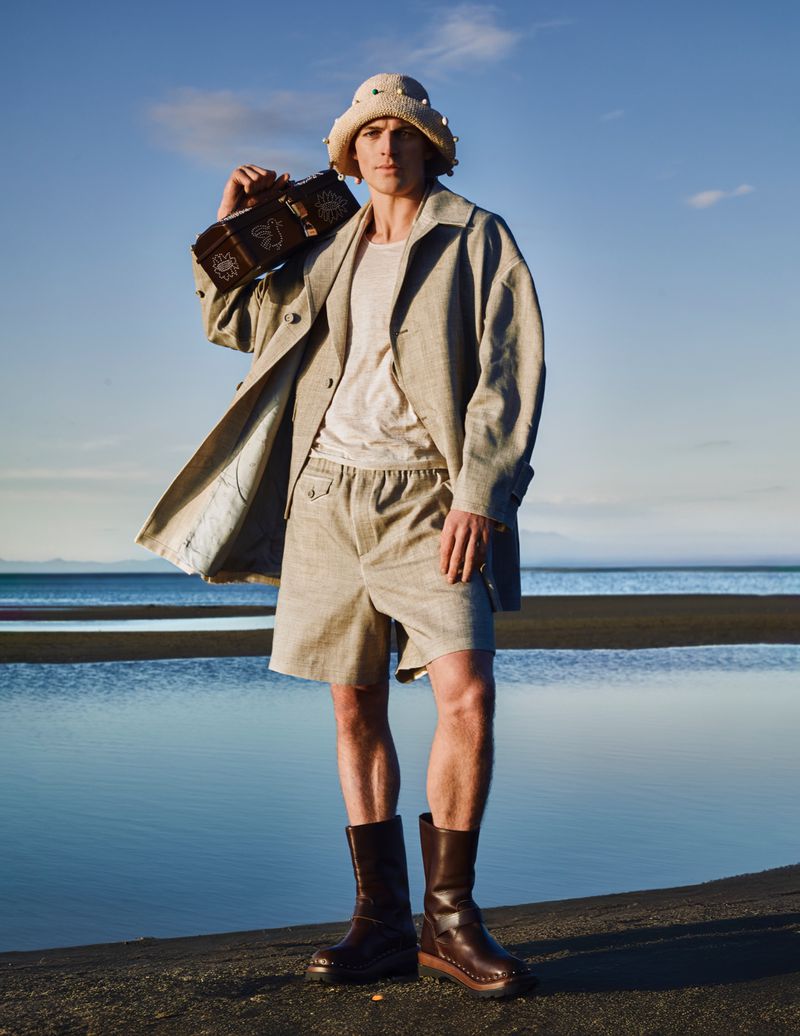 Man in oversized coat, shorts, and boots holds boombox on beach under blue sky, wearing embellished bucket hat in fashion style.