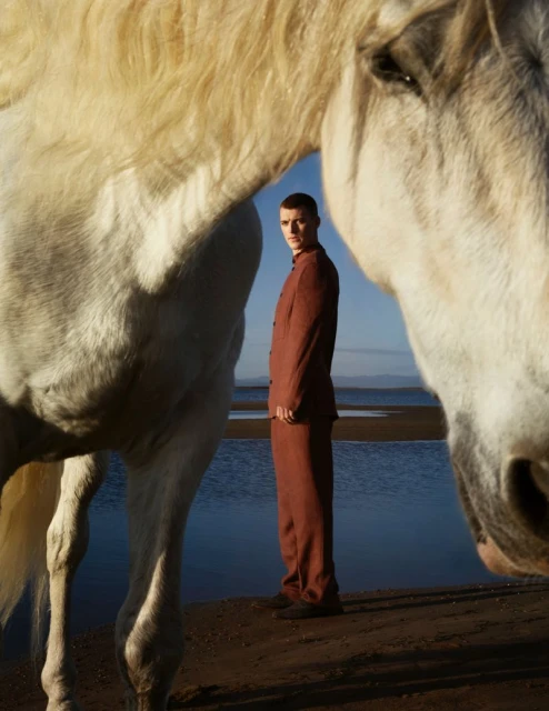 A man in a suit stands between the heads of two white horses on a beach with a sea and distant land under a clear sky.