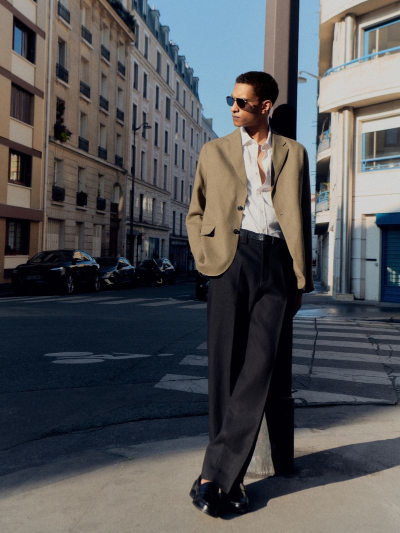 An adult male standing on a street corner, wearing a tan blazer, white shirt, black slacks, and sunglasses, posing during daylight hours.