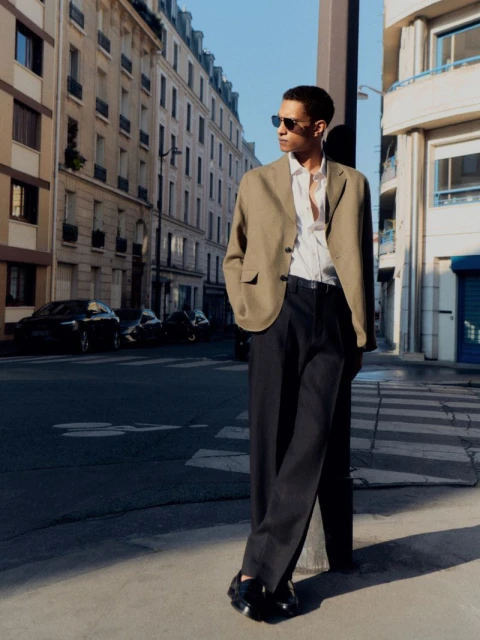 An adult male standing on a street corner, wearing a tan blazer, white shirt, black slacks, and sunglasses, posing during daylight hours.