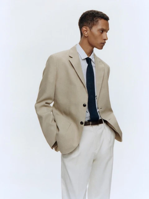 A dark-haired man poses in a beige suit with a blue tie against a white backdrop, showcasing a modern and professional fashion look.