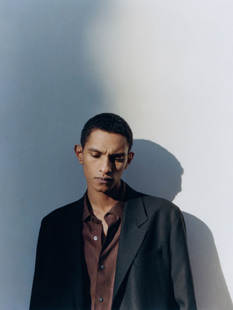 Man wearing a dark blazer and brown button-up shirt against a plain light wall with shadows.