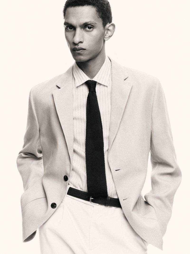 Man in a light suit, collared shirt, and dark knit tie poses against a light background in a studio. Fashion advertisement.