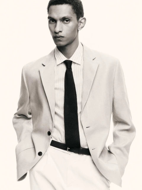 Man in a light suit, collared shirt, and dark knit tie poses against a light background in a studio. Fashion advertisement.