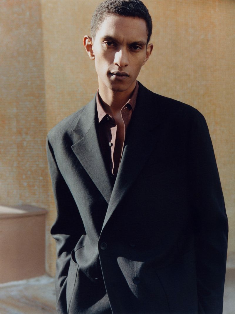 Dark-haired male model in a dark suit jacket and a buttoned brown shirt against a textured wall in an eye-level medium shot.