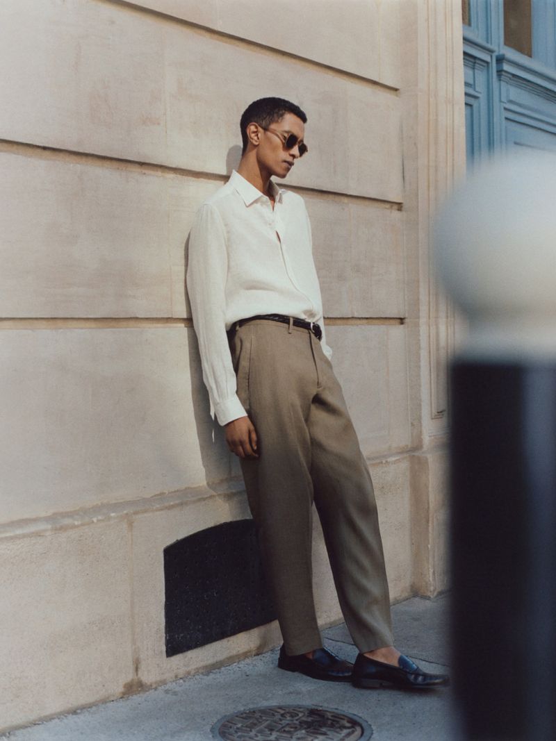 Man wearing a white shirt and khaki trousers leaning against a beige stone wall outdoors.