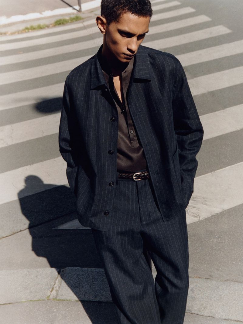 A young adult male walks on a crosswalk wearing a dark pinstripe suit with a brown collared shirt against an urban background.