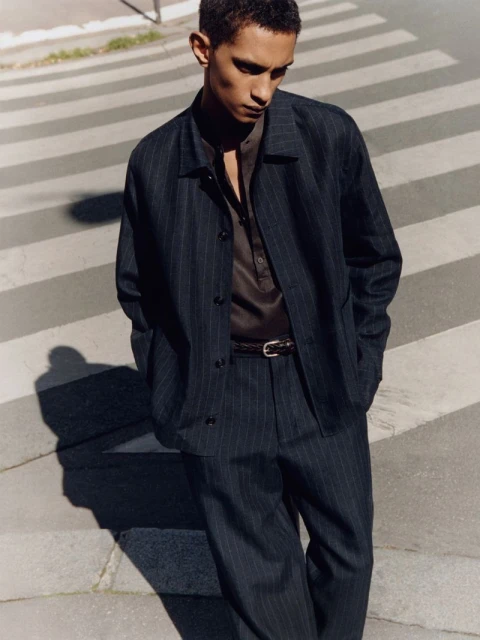A young adult male walks on a crosswalk wearing a dark pinstripe suit with a brown collared shirt against an urban background.