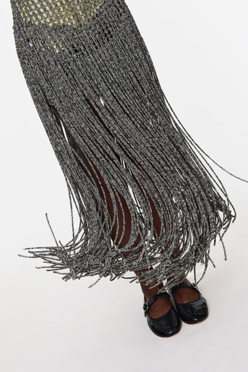 A partial shot of a person wearing a gray fringed dress with black patent leather Mary Jane shoes on a white background.
