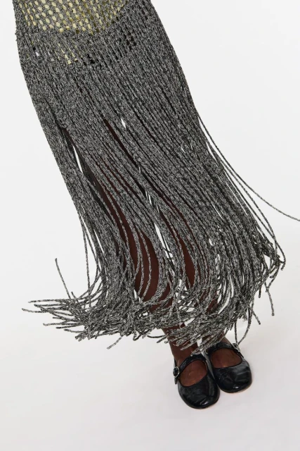 A partial shot of a person wearing a gray fringed dress with black patent leather Mary Jane shoes on a white background.