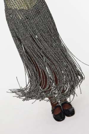 A partial shot of a person wearing a gray fringed dress with black patent leather Mary Jane shoes on a white background.
