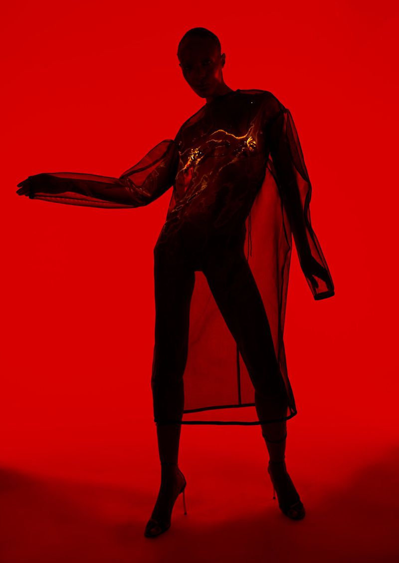 A model is silhouetted against a red backdrop, showcasing a transparent long-sleeved shirt and black pants with stiletto heels in a studio setting.