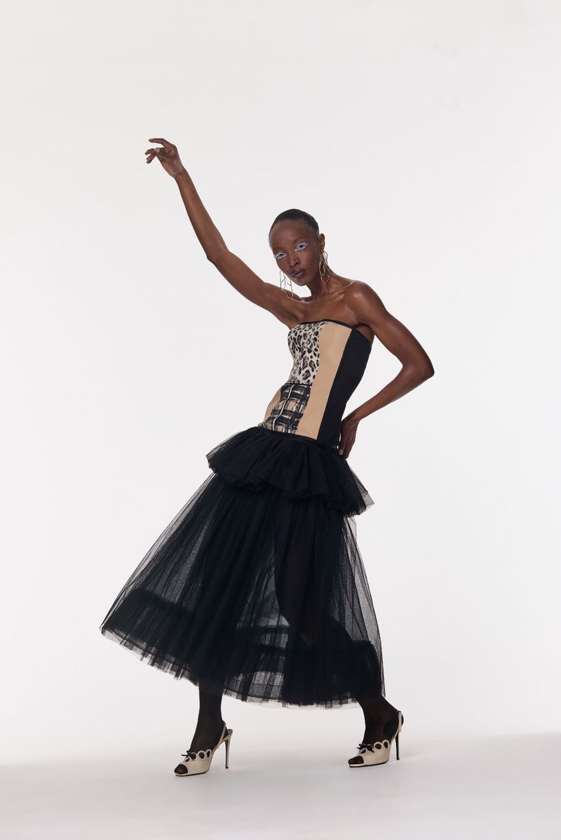 A slender, dark-skinned adult woman in a strapless corset top and tiered black tulle skirt strikes a graceful pose in a studio setting.