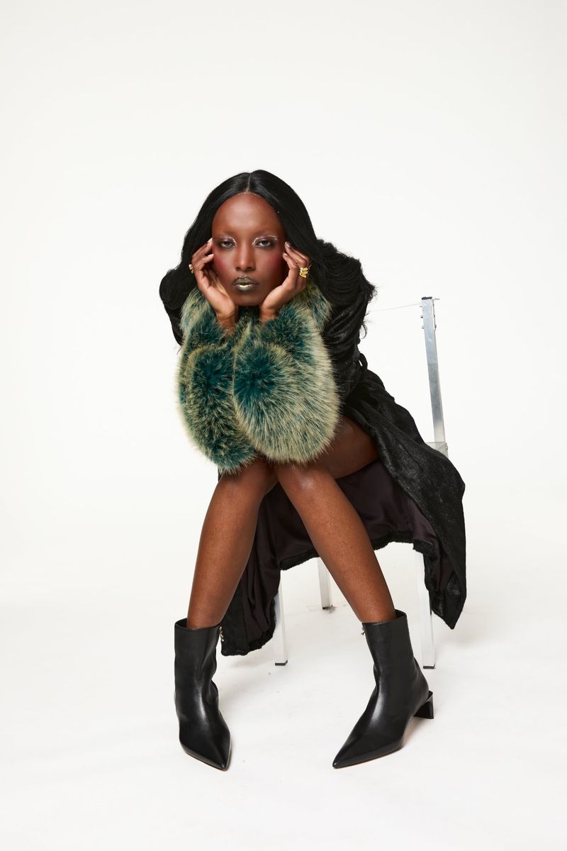 Elegant woman with dark hair, wearing black dress and boots, and faux fur sleeves, seated in a bright photo studio against a white backdrop.