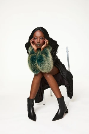 Elegant woman with dark hair, wearing black dress and boots, and faux fur sleeves, seated in a bright photo studio against a white backdrop.