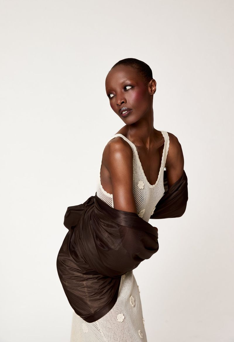An adult woman with short hair poses in a white dress with a brown garment draped over her shoulders against a plain white background.