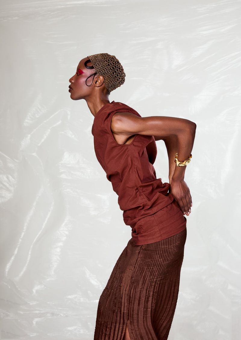 Model in studio wearing brown sleeveless top and brown pleated skirt, accessorized with gold bracelet and hair net on a white background.