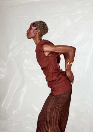 Model in studio wearing brown sleeveless top and brown pleated skirt, accessorized with gold bracelet and hair net on a white background.