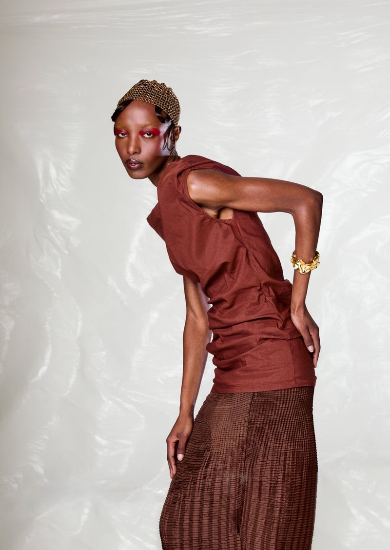 Dark-skinned model with red eye makeup, gold jewelry, and a brown dress poses in front of a light background in a studio shot.