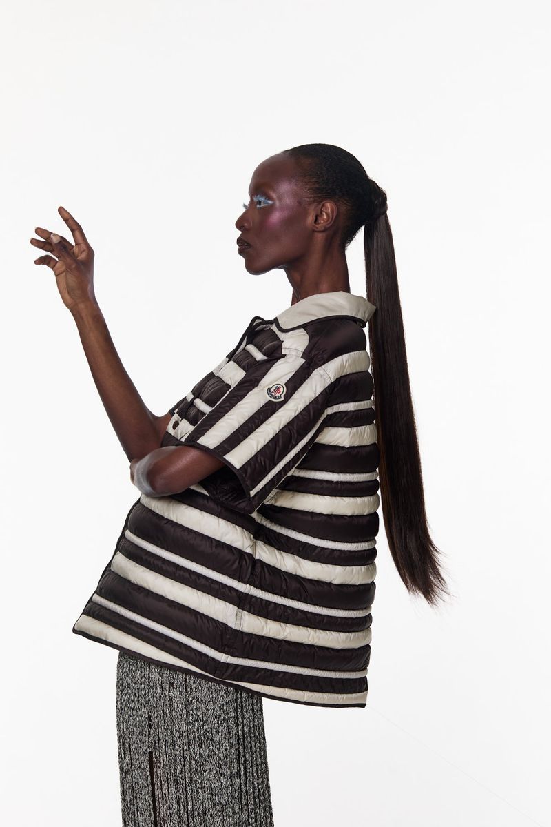 Black woman with long dark ponytail in striped puffer shirt raising hand against a white backdrop.