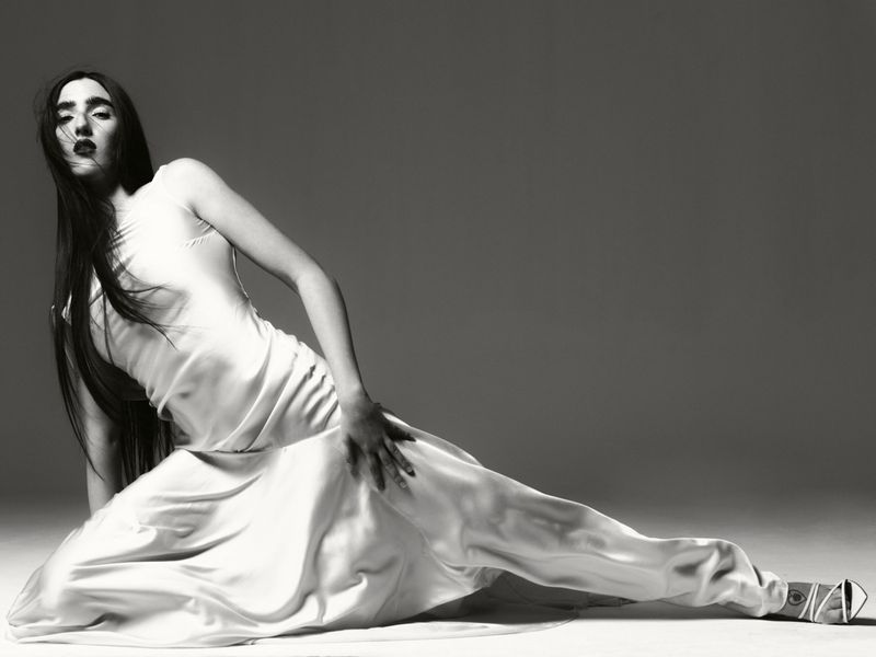 Elegant woman in a white slip dress reclining on a floor against a grey backdrop, showcasing a fashion-forward pose with dark lipstick and sleek hair.