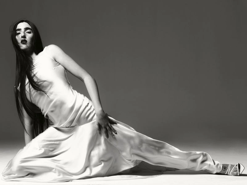 Elegant woman in a white slip dress reclining on a floor against a grey backdrop, showcasing a fashion-forward pose with dark lipstick and sleek hair.