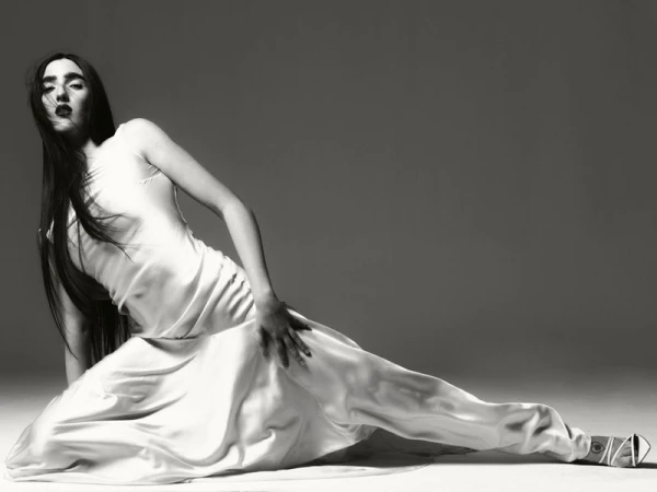 Elegant woman in a white slip dress reclining on a floor against a grey backdrop, showcasing a fashion-forward pose with dark lipstick and sleek hair.