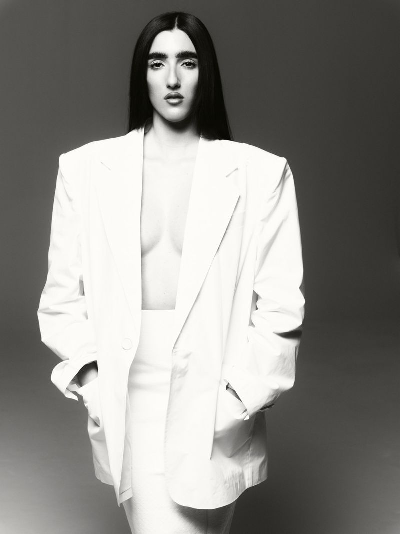Monochrome portrait of a woman in white suit with dark hair posing in studio, hands in pockets, confident expression, against grey backdrop.