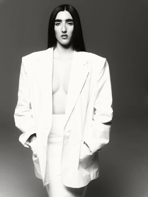 Monochrome portrait of a woman in white suit with dark hair posing in studio, hands in pockets, confident expression, against grey backdrop.