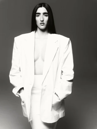 Monochrome portrait of a woman in white suit with dark hair posing in studio, hands in pockets, confident expression, against grey backdrop.
