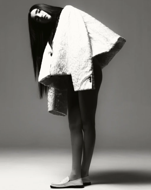 Monochrome fashion shot of a slender woman with long dark hair wearing a large white coat and loafers, standing in a studio.