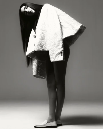 Monochrome fashion shot of a slender woman with long dark hair wearing a large white coat and loafers, standing in a studio.