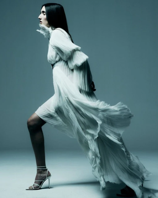 A woman in a long white flowing dress is walking and posing in a fashion shot with a light gradient background in a photo studio.