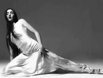 Monochrome fashion portrait of a woman with long dark hair wearing a white satin gown, posed dramatically on a light studio floor.