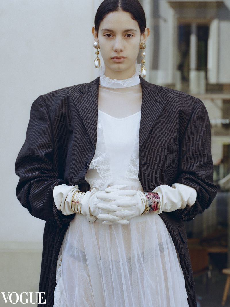 Portrait of a fashion model in white dress and dark blazer. The model has long dark hair, fair skin, and large dangling earrings.
