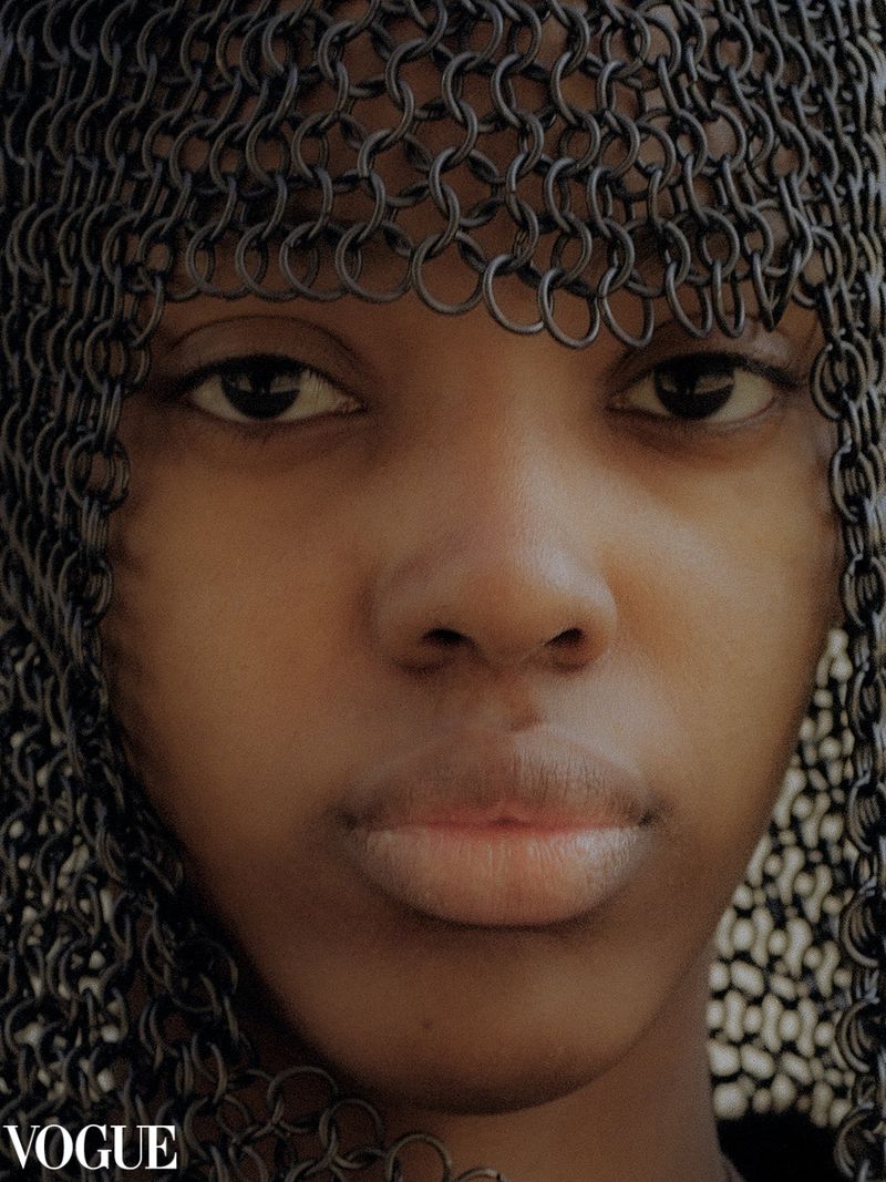 Close-up of a young person with dark hair wearing a chainmail head covering in a studio setting with the Vogue logo visible.